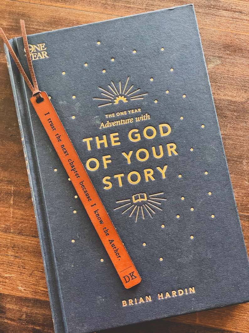 May include: A dark blue book with gold lettering that reads "The One Year Adventure with The God of Your Story."  The book has a brown leather bookmark with the text "I trust the next chapter because I know the Author." and the initials "DK".