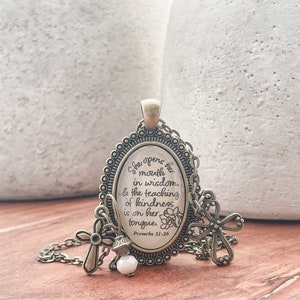 May include: A bronze necklace with a pendant featuring the quote "She opens her mouth in wisdom & the teaching of kindness is on her tongue. Proverbs 31:26". The pendant is surrounded by a decorative border and has a cross charm attached.