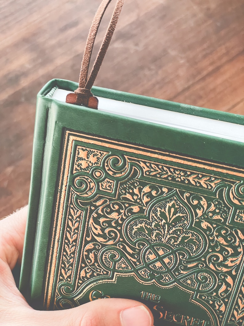 May include: A green hardcover journal with a gold embossed design and a brown leather bookmark. The title "The Secret Garden" is visible on the cover.