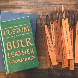 Puede incluir: Un conjunto de marcapáginas de cuero marrón con varios versículos bíblicos impresos en ellos. Los marcapáginas están dispuestos sobre una superficie de madera junto a dos libros con el texto "CUSTOM BULK LEATHER BOOKMARKS" en la portada.
