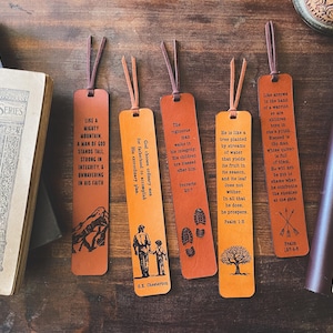 May include: Five leather bookmarks with inspirational quotes and illustrations. The bookmarks are various shades of orange and brown, with leather cords. The quotes are printed in black. A book and a journal are in the background.