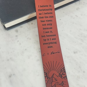 May include: A red leather bookmark with a quote by C.S. Lewis about Christianity. The quote reads: "I believe in Christianity as I believe that the sun has risen: not only because I see it, but because by it I see everything else." The bookmark features a sun rising over a mountain.