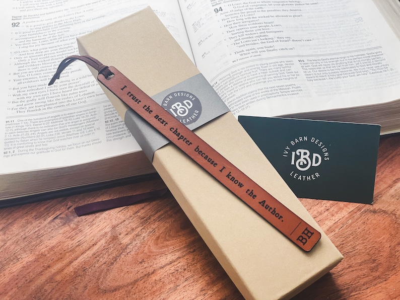 May include: A brown leather bookmark with the text "I trust the next chapter because I know the Author." and the initials "BH" engraved on it. The bookmark is packaged in a box with the logo "Ivy Barn Designs" and the text "Leather".
