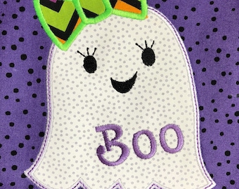 Puede incluir: Un fantasma blanco con un lazo verde y negro, costuras moradas y la palabra "BOO" bordada en la parte delantera. El fantasma está sobre un fondo morado con lunares negros.
