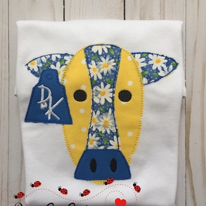 May include: White t-shirt with a yellow and blue cow applique. The cow has a daisy print on its head and a blue tag with the letter "R" on it. The shirt has a red heart and red ladybugs on the bottom.