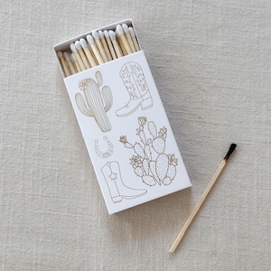 May include: A white box of matches with a gold cactus, cowboy boot, and horseshoe design. The box is open and a single match is lying next to it.