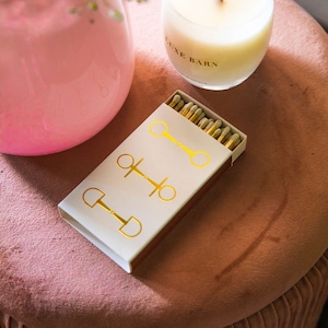 May include: A white box of matches with a gold equestrian bit design. The box is sitting on a pink velvet ottoman next to a clear glass candle with a wooden wick and the text "Luxe Barn" printed on the side.