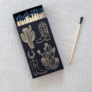 May include: A black matchbox with a gold design of cacti, cowboy boots, and a horseshoe. The matchbox is filled with wooden matches with black tips.