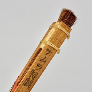 Puede incluir: Primer plano de un pincel de caligrafía japonesa. El pincel tiene un haz de cerdas marrones unidas a un mango de madera. El mango está hecho de finas láminas de madera de color claro sujetas con hilo. Se ve texto japonés.