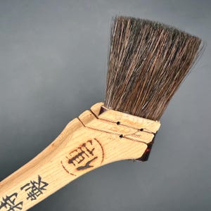 Pode incluir: Um pincel de madeira com cabo castanho claro e cerdas castanho escuro. O cabo tem caracteres japoneses. As cerdas são grossas e anguladas, adequadas para trabalhos de pintura detalhados.