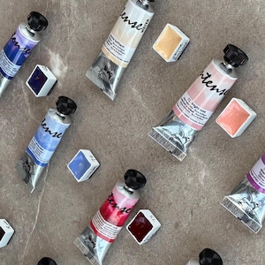 May include: A collection of Sennelier L'Aquarelle honey-based watercolor paint tubes and square pans in various colors including bright rose, lavender, and yellow. Each tube features a black cap and a label with the brand name Intense.