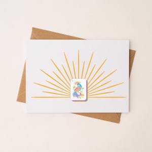 May include: White greeting card with a colorful illustration of a bird, set against a gold sunburst design. The card is placed on a brown envelope. The bird illustration features blue, pink, and yellow hues.
