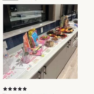 May include: A kitchen counter covered with a white runner featuring pink and blue graphic illustrations of cakes and the text "AH GIRLS". The runner is topped with various snack bags, bowls of food, and a pink patterned cup with metal utensils.