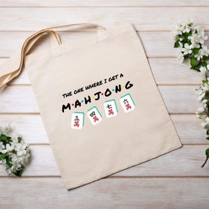 Puede incluir: Una bolsa de tela beige con el texto "The one where I get a Mahjong" y una fila de cinco fichas de mahjong con caracteres chinos.