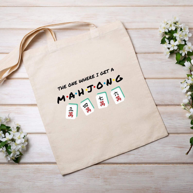 Puede incluir: Una bolsa de tela beige con el texto "The one where I get a Mahjong" y una fila de cinco fichas de mahjong con caracteres chinos.