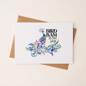 May include: A white greeting card with a colorful peacock illustration and the text "BIRD BAM IS MY JAM." The peacock design features blue, green, orange, and purple hues. The card is set on a brown envelope.