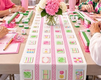 May include: A white table runner featuring a pink Greek key border and a repeating pattern of colorful mahjong tile illustrations. A glass vase filled with pink and white hydrangea flowers sits in the center of the table.