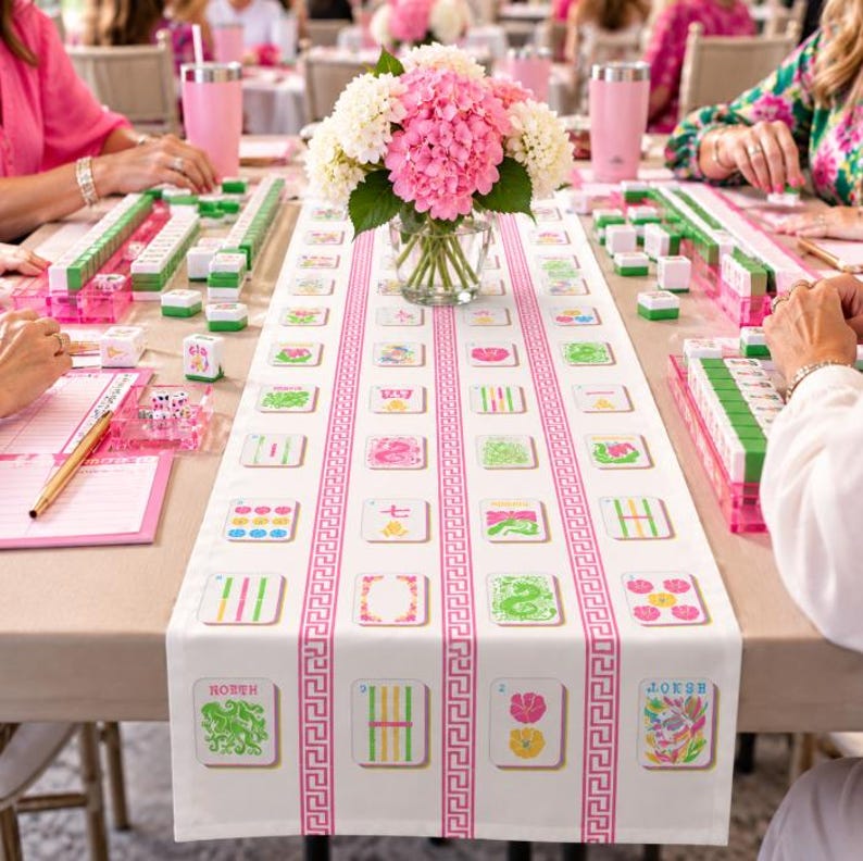 May include: A white table runner featuring a pink Greek key border and a repeating pattern of colourful mahjong tile illustrations. A glass vase filled with pink and white hydrangea flowers sits in the centre of the table.