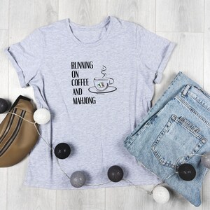 Puede incluir: Una camiseta gris con el texto "Running on coffee and mahjong" y una ilustraci&oacute;n en blanco y negro de una taza de caf&eacute; con vapor saliendo de ella.