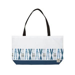 May include: A white tote bag with black shoulder straps featuring a repeating pattern of blue and white skis and ski poles across the lower section. The bottom of the bag is a solid dark blue band with a small rectangular label in the corner.
