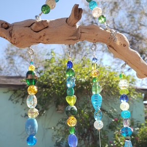 May include: A driftwood wind chime with strands of colorful glass beads in shades of green, blue, and yellow. The beads are strung on silver wire and hang from the driftwood.