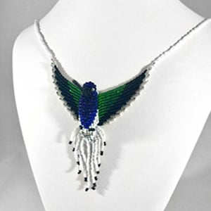 May include: A beaded hummingbird necklace displayed on a white mannequin. The necklace features a blue, green, and black bird design with white and black beaded accents. The wings are spread, and the necklace hangs from a silver chain.