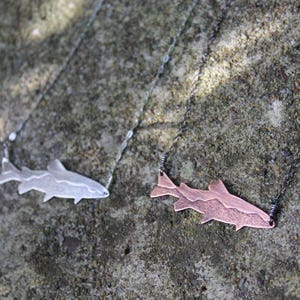 May include: Two metal fish pendant necklaces. One is silver-colored, the other is copper-colored. The fish pendants are connected to delicate chains. The necklaces are displayed on a textured, gray surface. The fish pendants are a unique jewelry item.