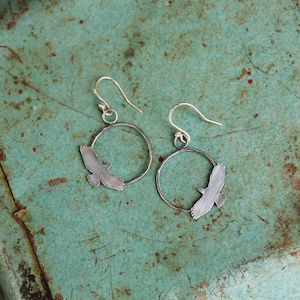 May include: A pair of silver earrings with a bird silhouette design. The earrings are circular and have a simple, minimalist style.