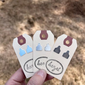 May include: Three pairs of earrings displayed on small cards with the word "bezel" printed on them. The earrings are silver, light blue, and dark gray.