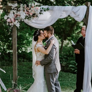 May include: A couple kisses under a white fabric canopy decorated with pink and white flowers. The couple is standing in front of a wooden archway. A person in a black suit is standing behind the couple, holding a book.