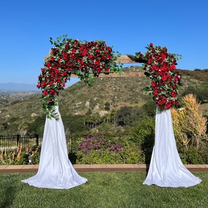 May include: A rustic wooden wedding arch decorated with red roses and greenery, with white fabric drapes on each side. The arch is set up outdoors against a backdrop of rolling hills.