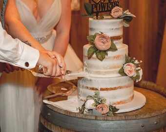 May include: A couple cuts a white wedding cake with a knife. The cake is decorated with flowers and has a sign that says "Mrs. Powers".