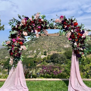 May include: A wooden wedding arch decorated with pink, red, and purple flowers and greenery. The arch is set up outdoors with a view of a green hillside in the background.