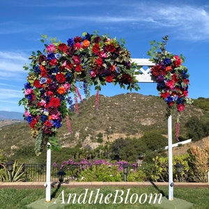 May include: A white wedding arch decorated with a large arrangement of colorful artificial flowers, including red, pink, purple, and blue roses. The arch is set up outdoors against a backdrop of green hills and a blue sky. The text "AndtheBloom" is visible in the foreground.