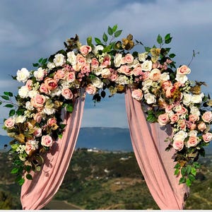 Op de afbeelding: Een roze bloemenboog gemaakt van kunstbloemen, met een roze stoffen achtergrond. De boog is versierd met roze, witte en perzikkleurige rozen, groen en andere bloemmotieven.