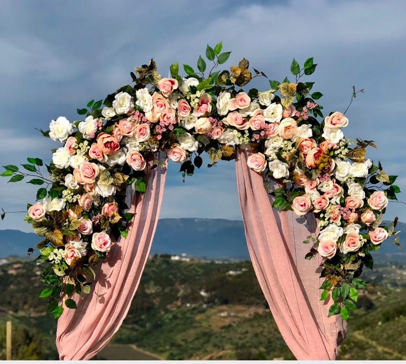 Op de afbeelding: Een roze bloemenboog gemaakt van kunstbloemen, met een roze stoffen achtergrond. De boog is versierd met roze, witte en perzikkleurige rozen, groen en andere bloemmotieven.