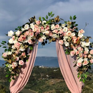 Op de afbeelding: Een bruiloftsboog versierd met roze en witte bloemen, groen en een roze stoffen achtergrond. De boog staat tegen een achtergrond van glooiende heuvels.