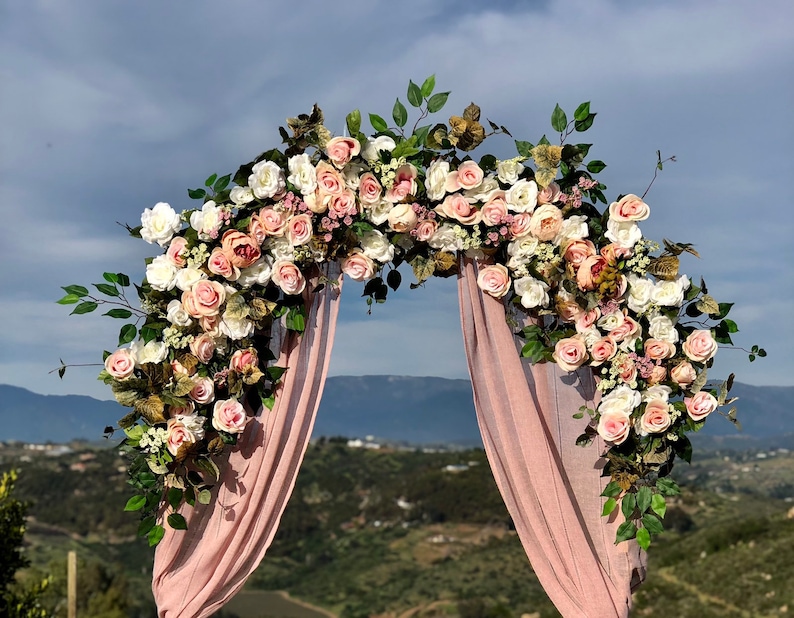 Op de afbeelding: Een bruiloftsboog versierd met roze en witte bloemen, groen en een roze stoffen achtergrond. De boog staat tegen een achtergrond van glooiende heuvels.