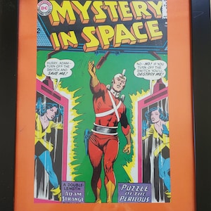 以下が含まれることがあります： 「Mystery in Space」と題された、額装されたヴィンテージコミックの表紙。表紙には、赤と白の衣装を着た人物が描かれており、「Mystery in Space」の文字は黄色です。コミックは黒いフレームにオレンジ色のマットで額装されています。