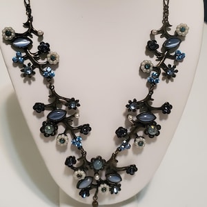 May include: A bronze-toned necklace with a floral design. The necklace features blue, white, and dark blue gemstones and faux pearls. A teardrop-shaped blue gem hangs from the center. The necklace is displayed on a white bust.