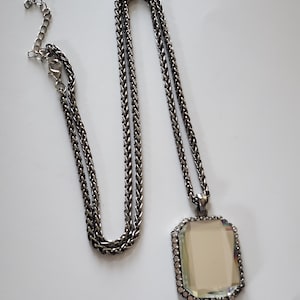 May include: A silver-toned necklace with a rectangular pendant. The pendant has a light-colored center and is framed with small, clear stones. The chain is a woven style, and the necklace has an adjustable length.