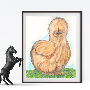 May include: A framed watercolor print of a Silkie chicken. The fluffy, light brown chicken is standing on green grass with small blue flowers. The artwork is in a black frame and is displayed on a white surface.