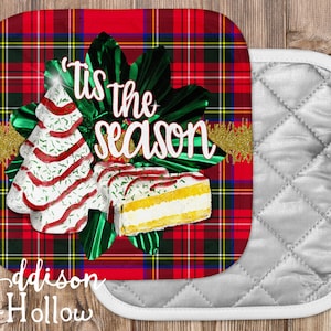 May include: A Christmas-themed pot holder with a red plaid pattern. The design features a Christmas tree and cake with the text "'Tis the season". The pot holder is paired with a quilted silver oven mitt.