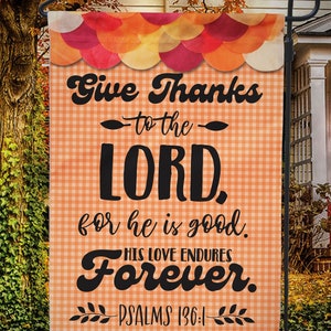 May include: A decorative garden flag with a fall theme. The flag features the text "Give Thanks to the LORD, for he is good. HIS LOVE ENDURES Forever. PSALMS 136:1" in black lettering on an orange gingham background. The top has a scalloped design.
