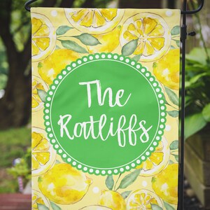 May include: A yellow garden flag with a green circle in the center. The circle has white polka dots around the edge and the text "The Ratliffs" in white. The background of the flag is a yellow watercolor pattern of lemons.