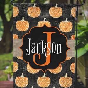 May include: A rectangular garden flag featuring a black background with a repeating pattern of orange pumpkins. A central black scalloped frame contains the name Jackson in white lettering with a large orange letter J. A burlap strip crosses the center.
