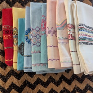 May include: A collection of ten folded kitchen towels in various pastel and vibrant colors including red, yellow, teal, light blue, pink, and white. Each towel features a unique, intricate embroidered geometric pattern in contrasting thread colors.