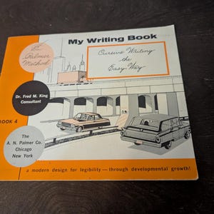 May include: A softcover instructional workbook titled My Writing Book, Cursive Writing the Easy Way, Book 4. The cover features an orange and white design with illustrations of cars on a highway, the Palmer Method logo, and text by Dr. Fred M. King.