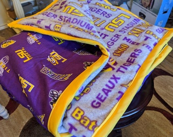 May include: A folded blanket featuring the LSU Tigers logo and text in purple and gold. The blanket has a purple interior with various logos and a gray exterior with text. The edges are trimmed with a yellow border.