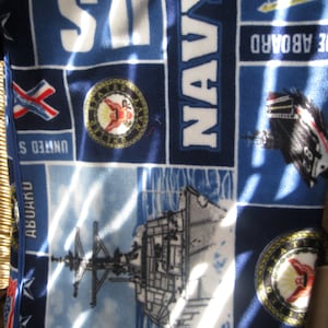 May include: A blue fleece blanket with a navy blue and white pattern. The blanket features a ship, the words "USS", "NAVY", and "Abukwa", and the seal of the United States Navy.